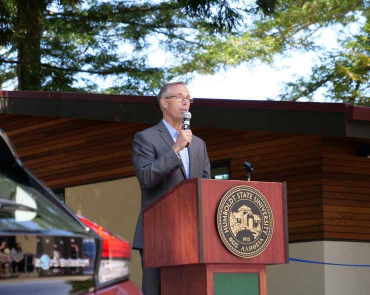 Jared Huffman speaks from a podium, between a zero-emission vehicle and the West Wing