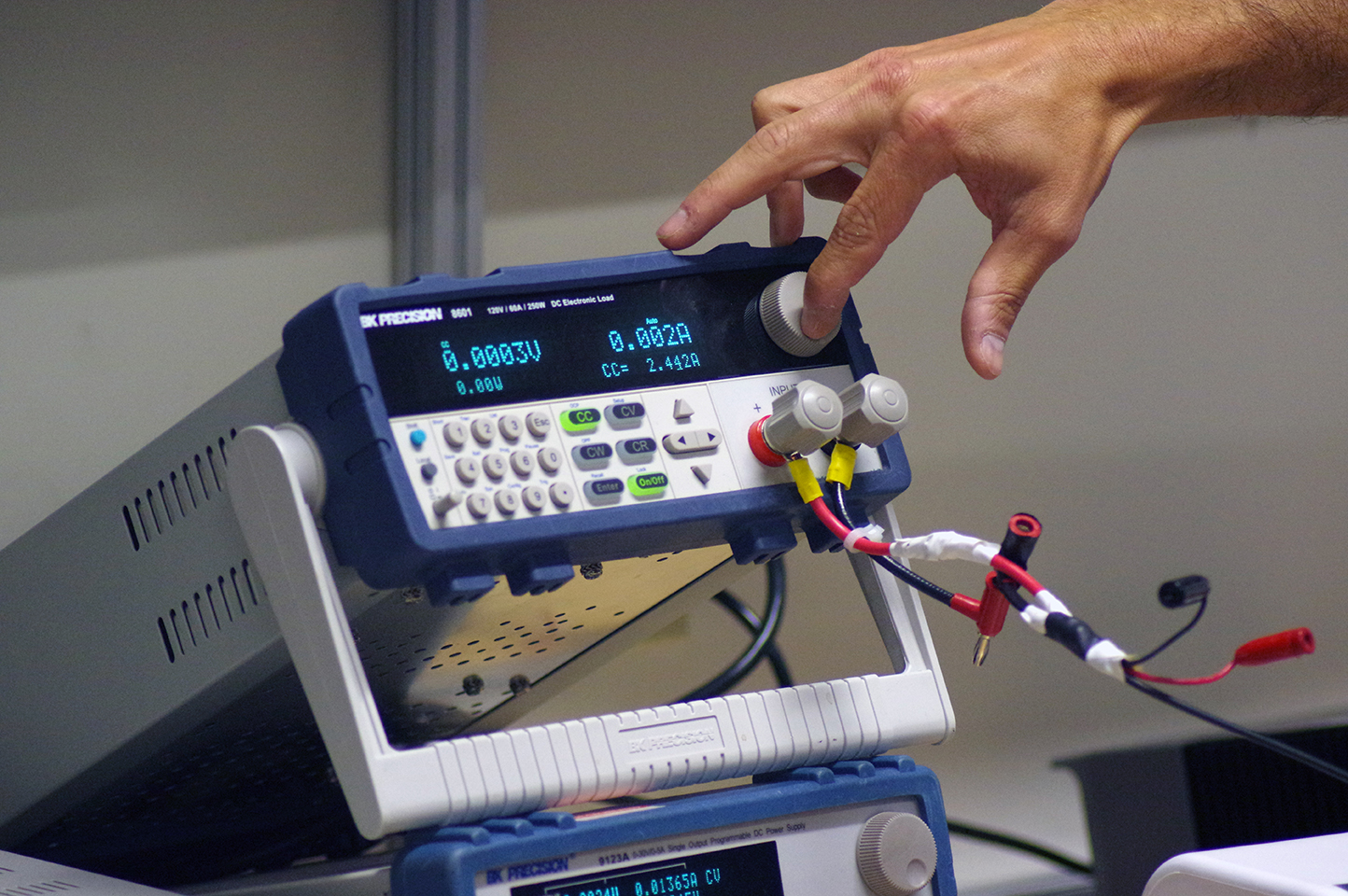 An extended hand turns the dial on an Electronic Load, which reads 0.0003V and 0.002A.