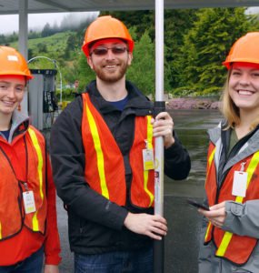 Three students in hardhats and orange vests just outside a gas station canopy.