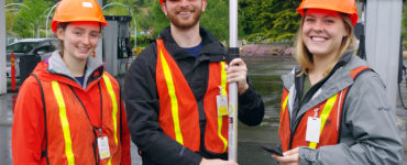Three students in hardhats and orange vests just outside a gas station canopy.