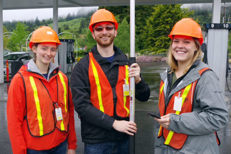 Three students in hardhats and orange vests just outside a gas station canopy.