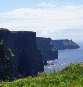 On a sunny day, steep, green-topped cliffs form a wave pattern against blue ocean
