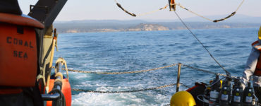 Two researchers sit near the stern with the shore receding in the distance