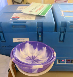 A blue, top-loading refrigerator with a manual on top sits behind nested purple and white plastic bowls and a tan plastic pitcher
