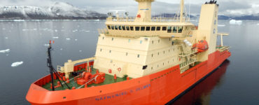 A ship with an orange base and cream decks is docked in a bay with snow-brushed mountains in the distance