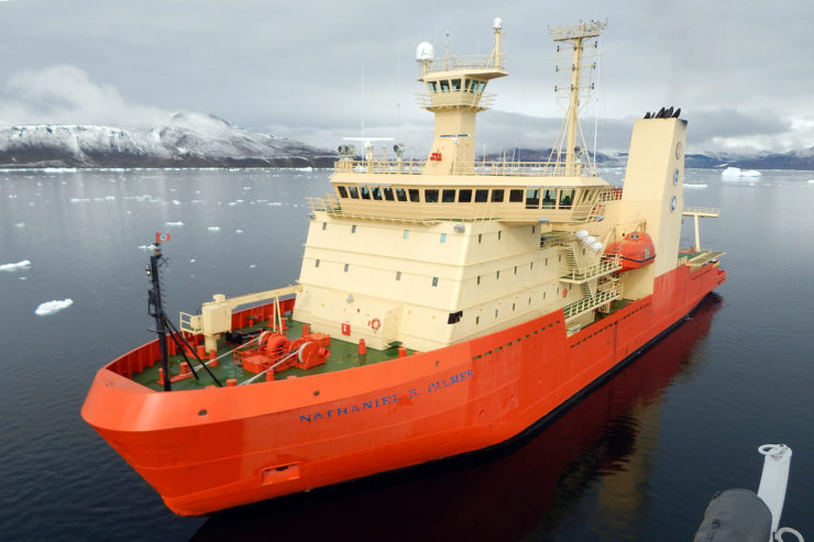 A ship with an orange base and cream decks is docked in a bay with snow-brushed mountains in the distance