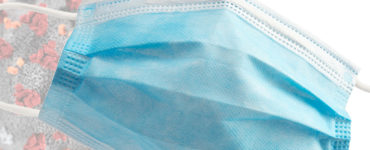 A blue surgical mask is superimposed over a graphic of a coronavirus
