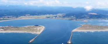 An aerial view of the entrance to Humboldt Bay, where a small boat enters between two jetties.