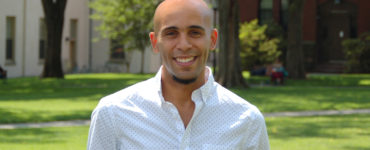 A smiling man in a white shirt stands on a grassy lawn
