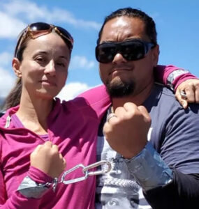 Two people stand with one arm around each other's shoulder and the other wrists locked together