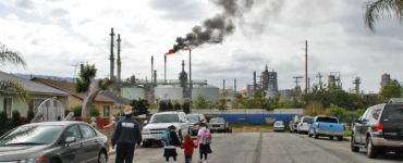 A family walks along a residential street, at the end of which is a refinery with a smoky plume