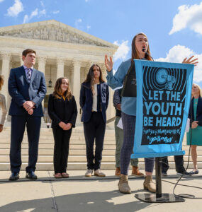 A young person speaks outside a courthouse, behind a banner that says "Let the youth be heard."