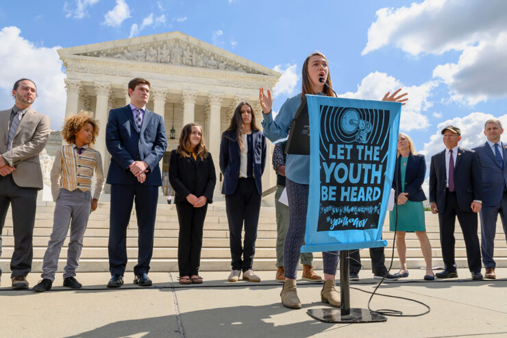 A young person speaks outside a courthouse, behind a banner that says "Let the youth be heard."